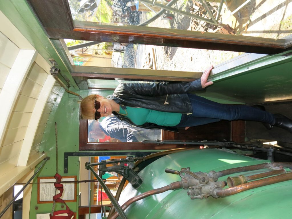 A woman wearing jeans and a leather coat is standing on a narrow wooden platform inside a steam tram. To her right is the side of a boiler and to her left are two windows. A steam tram is like a steam locomotive which travelled along streets. A wooden cab enclosed the entire locomotive, which features five windows along each side. Access to the cab is through doors from either a front or back platform. 